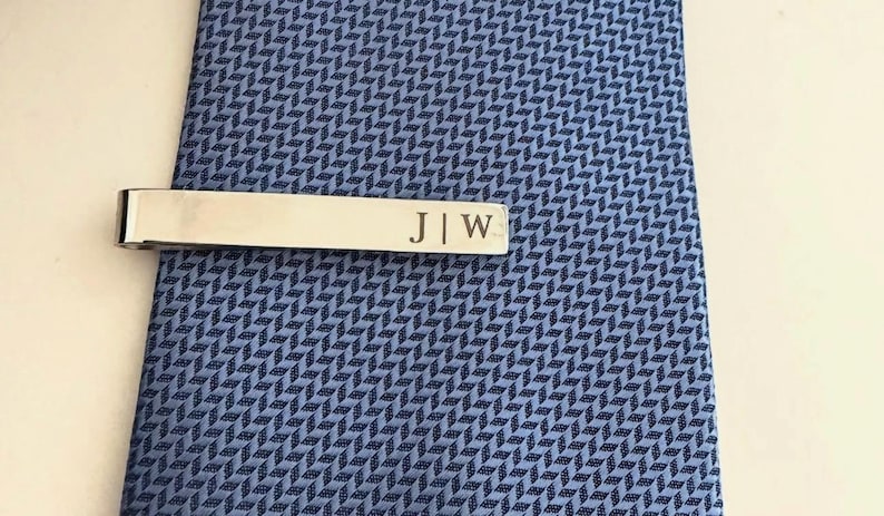 May include: A silver-toned metal tie bar featuring the engraved initials J | W, clipped onto a blue necktie with a repeating geometric herringbone pattern. The tie bar has a smooth, polished finish and is positioned horizontally across the fabric.