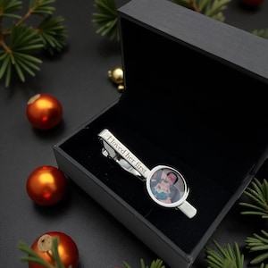 May include: A silver-toned metal tie clip featuring a circular photo insert of a person holding a child and the engraved text I loved her first, presented inside a black rectangular gift box with a hinged lid and a soft black interior lining.