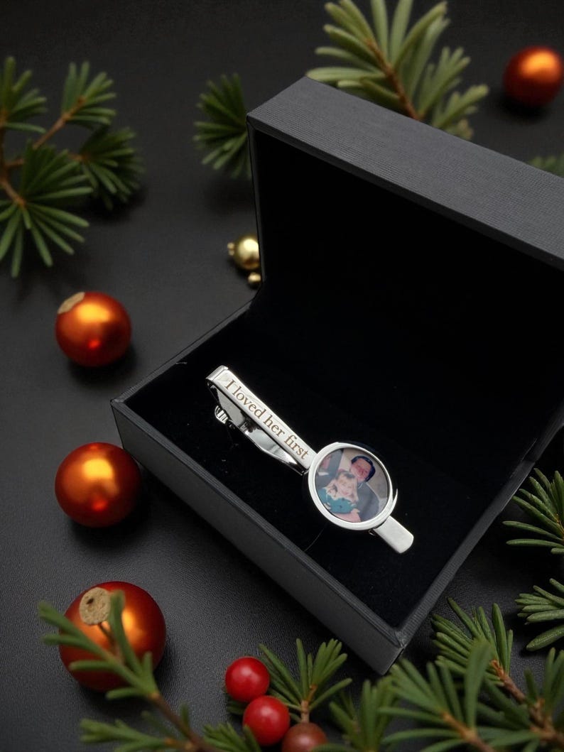 May include: A silver-toned metal tie clip featuring a circular photo insert of a person holding a child and the engraved text I loved her first, presented inside a black rectangular gift box with a hinged lid and a soft black interior lining.