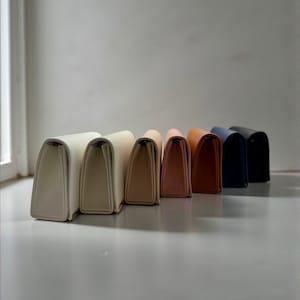 May include: A row of seven rectangular clutch purses in various colors, including cream, beige, tan, peach, orange-brown, navy blue, and black. The purses are arranged in a line, with their tops facing the viewer.