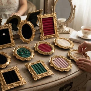 May include: A collection of ornate, gold-colored jewelry display trays and frames. The trays feature velvet inserts in various colors, including red, green, and pink. Some frames hold rings and earrings, while others are empty.