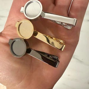 May include: Three tie clips with circular bezels for customization, featuring a polished silver-tone finish, a bright gold-tone finish, and a dark gunmetal-tone finish. Each clip has a spring-loaded mechanism for securing to a necktie.