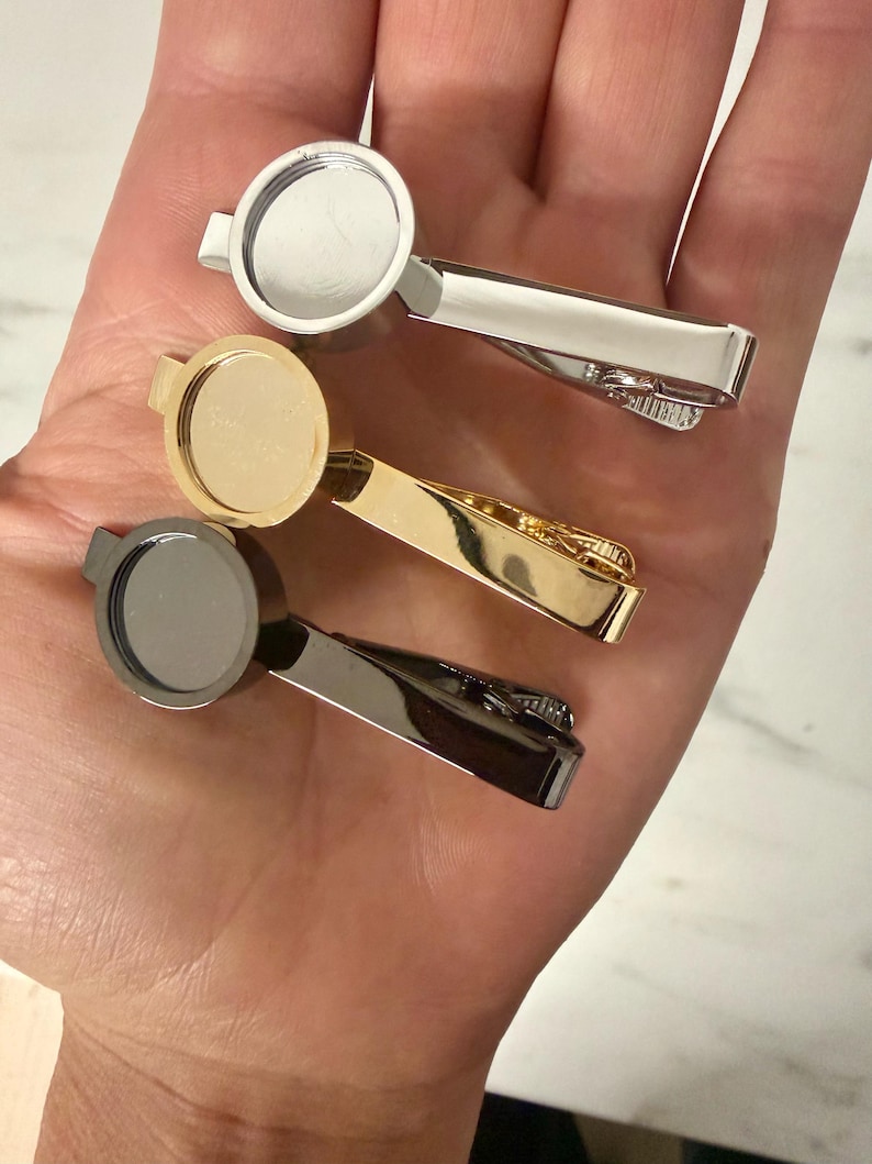 May include: Three tie clips with circular bezels for customization, featuring a polished silver-tone finish, a bright gold-tone finish, and a dark gunmetal-tone finish. Each clip has a spring-loaded mechanism for securing to a necktie.