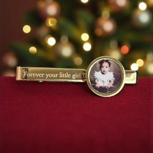 May include: A gold-toned tie clip featuring a rectangular bar engraved with the text "Forever your little girl" and a circular frame containing a small, circular photograph of a young child wearing a white ruffled top.
