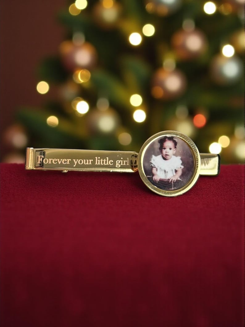 May include: A gold-toned tie clip featuring a rectangular bar engraved with the text "Forever your little girl" and a circular frame containing a small, circular photograph of a young child wearing a white ruffled top.