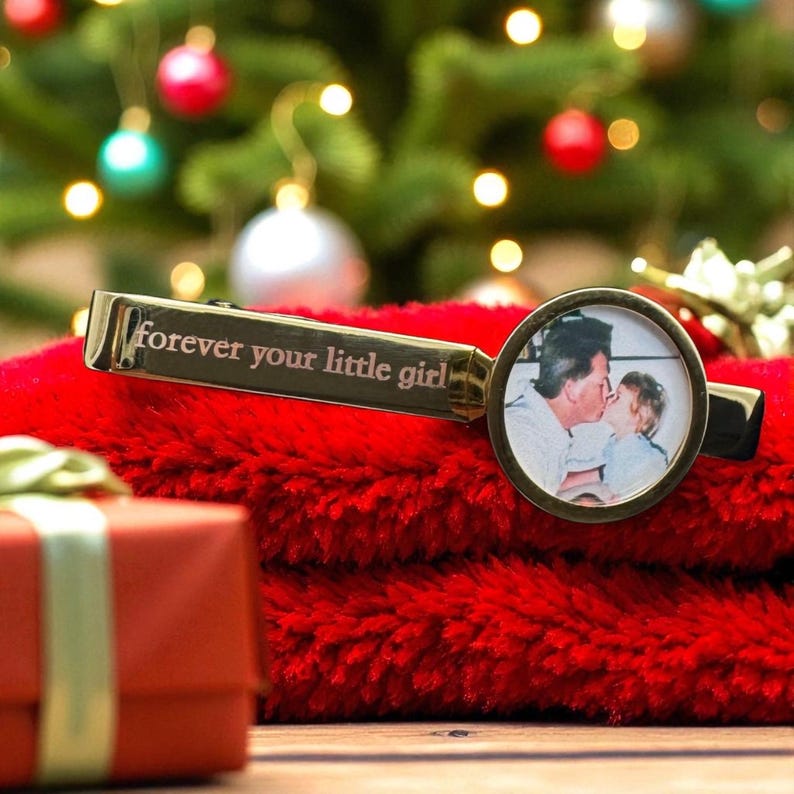 May include: A gold-toned metal tie clip featuring an engraved inscription that reads "forever your little girl" and a circular frame containing a black and white photograph of an adult kissing a child on the cheek.