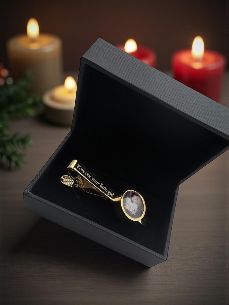 May include: A gold-toned tie clip featuring a small oval photo insert and the engraved text "Forever your little girl" on the bar, presented inside a black rectangular gift box with a black velvet interior lining.