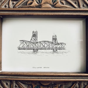 May include: A framed black and white sketch of the Stillwater Bridge. The bridge is a detailed drawing with two tall towers and a curved central span. The frame is a decorative, ornate, gold-toned wood.