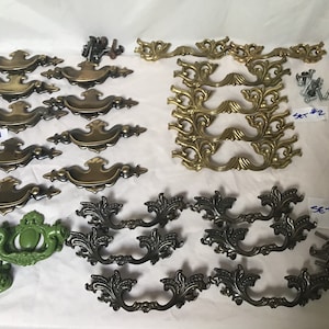 May include: A collection of ten sets of decorative cabinet hardware. The sets include ornate brass, black, and green drawer pulls. The pulls are in various styles, including floral and scroll designs. The image also includes screws for installation.