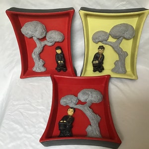 Puede incluir: Tres placas de pared decorativas con fondo rojo, amarillo y rojo. Cada placa presenta un árbol estilizado gris con una persona vestida de negro de pie junto a él.