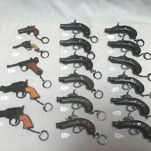 May include: A collection of 16 toy gun keychains, each with a different design and number. The keychains are made of metal and have a black finish. Some of the keychains have wooden handles. The keychains are arranged in rows on a white surface.