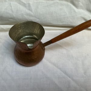 May include: A hammered copper coffee pot featuring a wide, circular opening and a pouring spout. The interior has a silver-toned metallic finish. A long, slender, light brown wooden handle is attached to the side of the pot with a metal rivet.