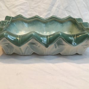 Peut inclure: Un pot de fleurs rectangulaire en céramique avec un bord festonné. Le pot est vert clair avec une finition brillante. L'intérieur est d'une couleur plus claire, presque blanche. Le pot est destiné à un usage intérieur et peut contenir de petites plantes ou fleurs.