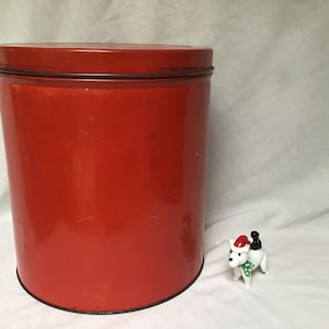 May include: A bright red metal tin with a smooth, shiny surface. The tin is cylindrical and has a lid.