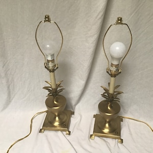 May include: A pair of gold pineapple table lamps with white lampshades and clear light bulbs. The lamps have a square base and a pineapple-shaped design.
