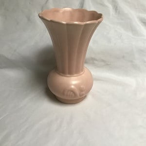 May include: A pale pink ceramic vase with a fluted design and a raised floral motif. The vase has a rounded base and a flared opening.