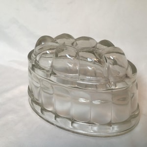 May include: A clear glass jelly mould with a rounded rectangular shape. The mould features vertical ridges along the sides and a decorative, rounded top. The glass is transparent, allowing light to pass through.