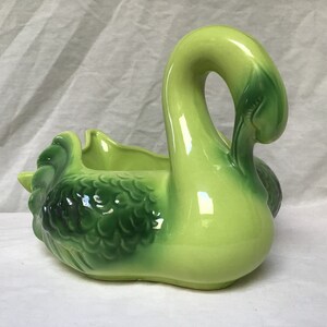 May include: A green ceramic swan planter with a glossy finish. The swan has a detailed design of feathers and a wide opening for planting.