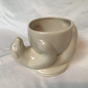 May include: A cream-coloured ceramic seal-shaped planter. The seal is depicted lying down, with a round opening for planting on its back. The planter is suitable for small plants or succulents and is made of ceramic.