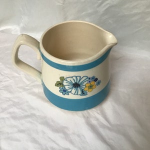 Op de afbeelding: Een witte keramische kan met een blauwe band en een bloemenontwerp. De kan heeft een handvat en een tuit.