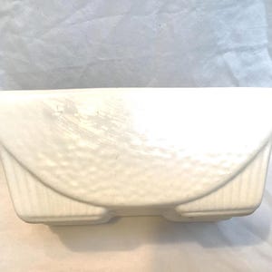 Könnte beinhalten: Rechteckiger, cremefarbener Keramik-Blumentopf mit strukturierter, gewölbter Front. Der Pflanztopf hat ein geriffeltes Design an den Seiten und eine glatte, abgerundete Oberseite. Für den Innen- oder Außenbereich.