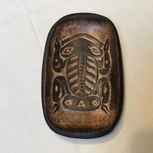 May include: A dark brown, oval leather tray featuring a carved frog design. The frog is centrally positioned and has detailed features. The tray has a distressed, aged appearance.