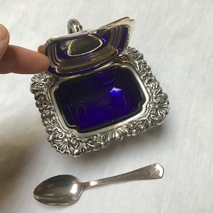 Könnte beinhalten: Silberne Salzdose mit blauer Glasauskleidung und passendem Löffel. Der Deckel ist geöffnet und die Salzdose hat ein verziertes Design.