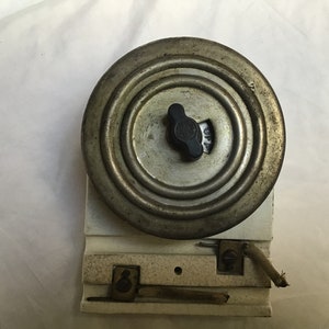May include: A vintage, round, metal electrical meter with a black knob and a white porcelain base. The meter has a circular dial with numbers and a needle.