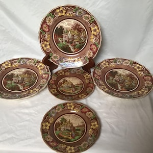 May include: Set of five brown and cream ceramic plates with floral accents and a cottage scene in the centre.