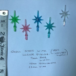 May include: Five small, iridescent star-shaped Christmas tree toppers with a lustre finish. The stars are in shades of green, pink, blue, and clear. The text below the stars reads: "Small stars with lustre finish. (Very limited supply) 4mm base. 45mm total length. 23mm wide."