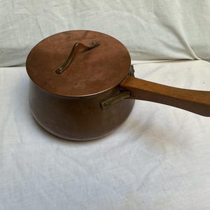 Peut inclure: Une casserole ronde en cuivre avec un couvercle assorti et un long manche en bois. La casserole présente une finition en cuivre métallique, une fixation de manche couleur laiton et une poignée en bois courbée fixée par des rivets métalliques.