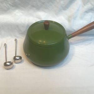 Könnte beinhalten: Ein grüner Emaille-Fondue-Topf mit Holzgriff und Deckelknopf, begleitet von zwei kleinen silbernen Schöpflöffeln. Der Topf hat eine runde Form und einen Deckel. Die Schöpflöffel sind klein und rund.