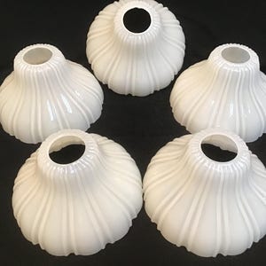 Op de afbeelding: Vijf witte glazen lampenkappen met een geribbeld, tulpachtig ontwerp. De kappen hebben een brede, afgeronde basis en een smallere, cirkelvormige opening aan de bovenkant. De kappen zijn in een cirkelvormig patroon tegen een donkere achtergrond geplaatst.