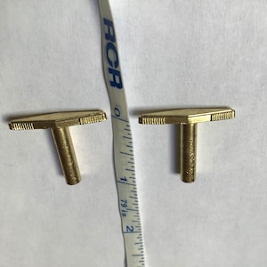 May include: Two gold-colored T-shaped keys with a diamond-shaped head and a small groove around the edge. The keys are about 1 inch long and are sitting on a white surface.