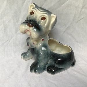 Może przedstawiać: Ceramiczna doniczka w kształcie szarego i białego psa z czerwono-białym kołnierzem w kropki. Pies ma smutny wyraz twarzy i duże oczy.