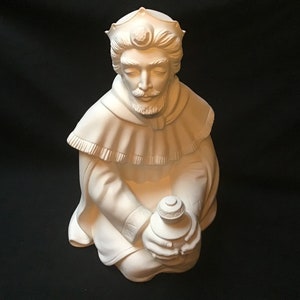 May include: White ceramic figurine of a kneeling king wearing a crown and holding a small jar. The king has a beard and is dressed in a robe with a fringed collar.