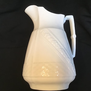 May include: A white ceramic jug with a decorative handle and spout. The jug features embossed diagonal and horizontal patterns. The handle has a bamboo-like design. The jug is a light, off-white colour.