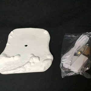 Może przedstawiać: Biała ceramiczna podstawa z fakturowaną powierzchnią i otworem w środku. Oddzielny plastikowy worek zawiera biały przewód, białą wtyczkę i małą brązową żarówkę.