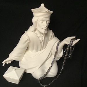 May include: A white ceramic bust of a crowned figure holding a rosary with silver beads. A small, white, lidded box is to the left. The figure has a beard and wears a robe. The background is black.
