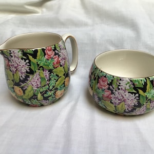 May include: A vintage floral creamer and sugar bowl set. The set features a black background with a pattern of pink, purple, and yellow flowers with green leaves. The rims are trimmed with gold. The creamer has a handle and spout.