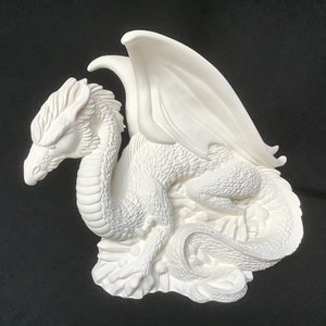 Peut inclure: Figurines de dragon en céramique blanche avec des ailes déployées, assise sur une base rocheuse. Le dragon a des écailles détaillées et une bouche fermée.