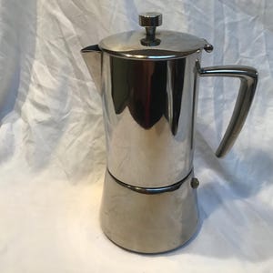 May include: A stainless steel stovetop espresso maker with a lid, handle, and spout. The coffee maker has a cylindrical body and a rounded base. The handle is a curved, solid piece of metal. The coffee maker is designed to brew espresso.