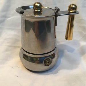May include: A stainless steel stovetop espresso maker with a gold-coloured handle and lid knob. The coffee pot has a round base and a cylindrical body. The handle and lid knob are a shiny gold colour. The espresso maker is designed for brewing coffee.