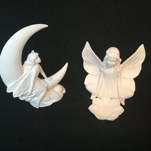 Peut inclure: Deux figurines de fée en céramique blanche. Une fée est assise sur un croissant de lune, l'autre tient une fleur.