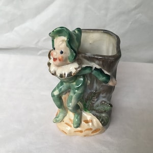 May include: A ceramic planter shaped like a green elf holding a brown tree trunk. The elf has a white ruff and a green hat. The planter has a square opening at the top.