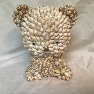May include: A small bear sculpture made entirely of seashells. The bear has a white and brown shell body with two black eyes.