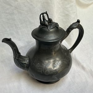 May include: A dark grey metal teapot featuring an ornate handle and a curved spout. The lid is topped with a sculpted floral finial. The body of the teapot is decorated with delicate, etched swirling patterns and leaf motifs along the base of the spout.