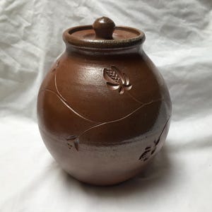 May include: A round, brown ceramic jar with a matching lid featuring a small knob handle. The exterior surface of the jar is decorated with carved, stylised floral or leaf patterns and thin, curved lines etched into the clay body.