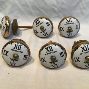 Może przedstawiać: Zestaw gałek do szafek z tarczą zegara. Każda gałka ma białą tarczę z czarnymi rzymskimi cyframi, napisem "POTTER-STONE" oraz złotą obwódką i okuciami. Gałki są zaprojektowane tak, aby przypominały antyczne tarcze zegarowe.