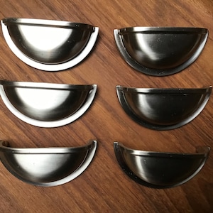 May include: Six silver cup-shaped cabinet pulls with a rounded edge. The pulls are arranged in two rows of three on a wooden surface.